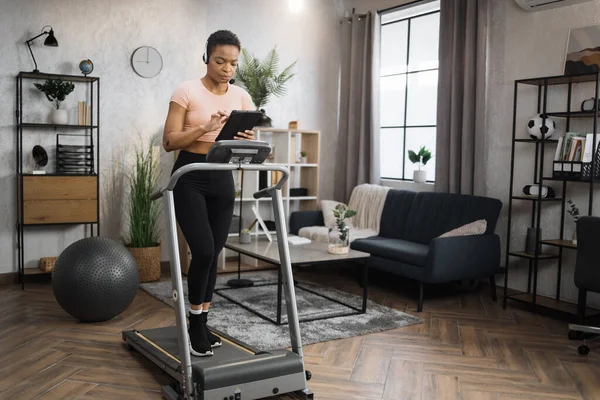 Side view of african woman in sport clothes using headset working on tablet computer while doing cardio training on treadmill at morning at home gym. Concept of sport, health care, business.