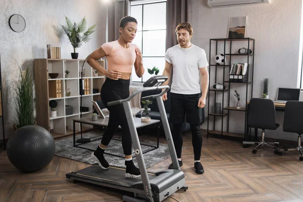 Home fitness workout sporty woman and man training on running track. Portrait of focused young african female wearing sportswear and using treadmill and her trainer bearded caucasian male.