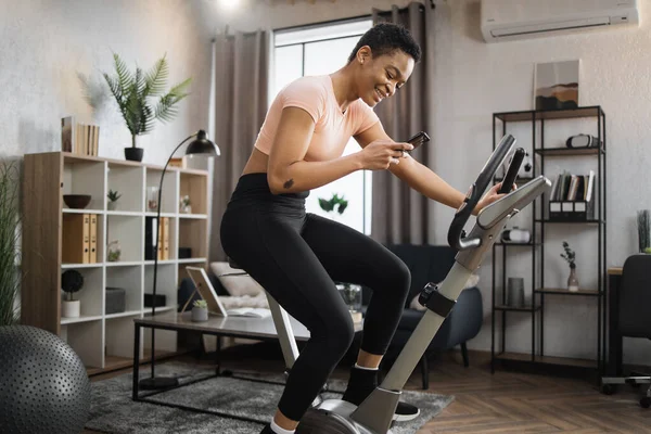 Smiling beautiful african american sports woman in sportswear writing message on social media, networks while cycling bike at home on background of light living room.
