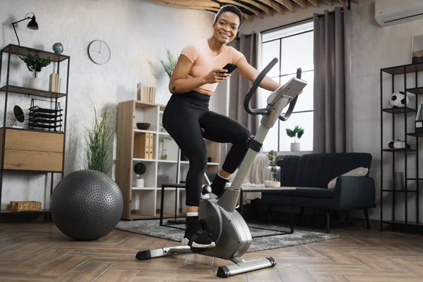 Low angle view of smiling beautiful african american sports woman in sportswear writing message on social media, networks while cycling bike looking at camera on background of light living room.