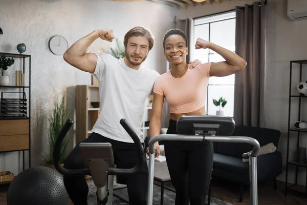 Smiling african woman running on treadmill and caucasian man training on exercise bike showing biceps looking at camera. Young couple doing cardio on stationary bike and treadmill.