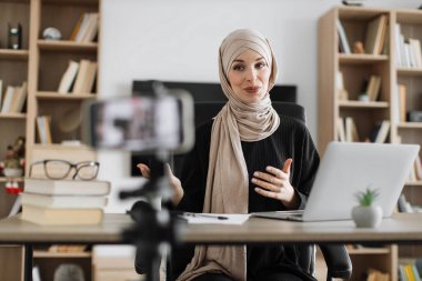 Charming muslim woman in hijab talking and gesturing while recording video on modern phone. Female blogger sitting at home and doing live stream.