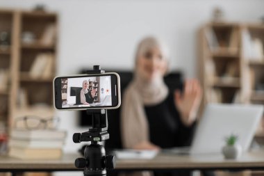 Blur background of pretty muslim woman with headscarf sitting at desk and filming video blog. Focus on modern digital phone fixed on tripod.