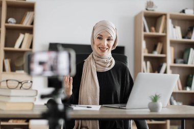 Smiling arab woman in beige hijab sitting at desk with books, notes and laptop recording video blog talking with followers. Female tutor working at home during distance learning.