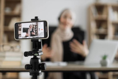 Charming muslim woman in hijab and headset talking and gesturing while recording video on modern phone. Female blogger sitting at home and doing live stream.