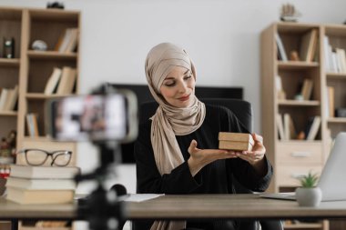 Pretty muslim woman in casual wear and hijab recording video on camera while unpacking gift boxes. Female blogger sharing her emotions with her subscribers in social networks.