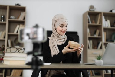 Pretty muslim woman in casual wear and hijab recording video on camera while unpacking gift boxes. Female blogger sharing her emotions with her subscribers in social networks.