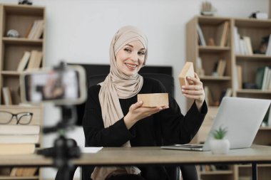 Happy muslim female blogger unpacking gift boxes presents from companies and doing live streaming. Arab woman using smart phone camera for creating content.