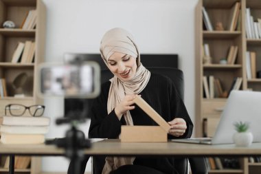 Happy muslim female blogger unpacking gift boxes presents from companies and doing live streaming. Arab woman using smart phone camera for creating content.
