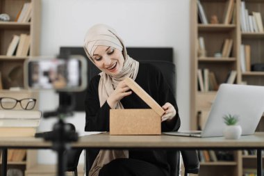 Happy muslim female blogger unpacking gift boxes presents from companies and doing live streaming. Arab woman using smart phone camera for creating content.