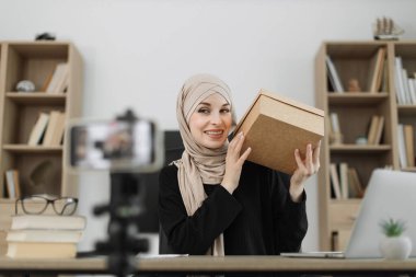 Excited smiling muslim woman in casual wear and hijab recording video on camera while unpacking gift boxes. Female blogger sharing her emotions with her subscribers in social networks.