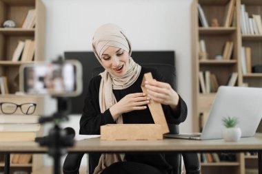 Happy muslim female blogger unpacking gift boxes presents from companies and doing live streaming. Arab woman using smart phone camera for creating content.