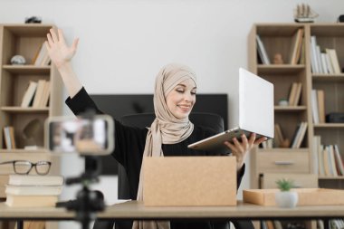 Pleasant arab woman filming video on modern phone camera while opening parcel box with new wireless laptop. Concept of people, technology and blogging.