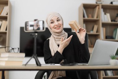 Pretty muslim woman in casual wear and hijab recording video on camera while unpacking gift boxes. Female blogger sharing her emotions with her subscribers in social networks.