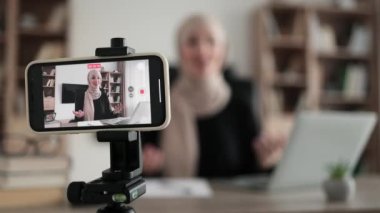 Blur background of pretty muslim woman with headscarf sitting at desk and filming video blog. Focus on modern digital phone fixed on tripod.