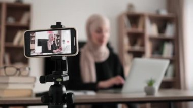 Confident muslim female in headscarf sitting at desk working on laptop and recording video on digital smartphone. Concept of freelance and modern technology.
