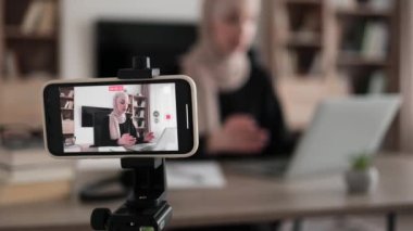 Confident muslim female in headscarf sitting at desk working on laptop and recording video on digital smartphone. Concept of freelance and modern technology.