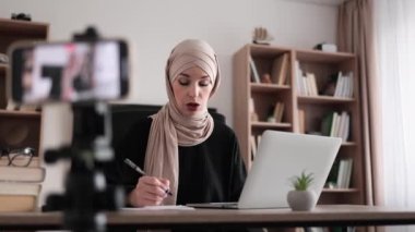 Cheerful young muslim woman in hijab recording creative video blog for her social networks, media for followers while working with laptop and writing report.
