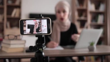 Focus on screen of phone of cheerful young muslim woman in hijab recording creative video blog for her social networks, media for followers while working with laptop and writing report.