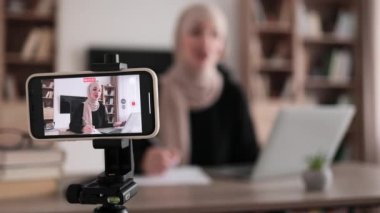 Focus on screen of phone of cheerful young muslim woman in hijab recording creative video blog for her social networks, media for followers while working with laptop and writing report.