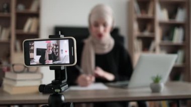 Focus on screen of phone of cheerful young muslim woman in hijab recording creative video blog for her social networks, media for followers while working with laptop and writing report.