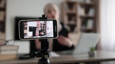 Focus on screen of phone of happy arab woman sitting at desk with books, notes and laptop talking with followers. Female tutor working at home during distance learning and showing thumb up.