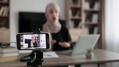 Focus on screen of phone of happy arab woman sitting at desk with books, notes and laptop talking with followers. Female tutor working at home during distance learning and showing sign ok.