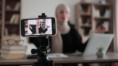 Blurred relaxed young muslim woman in casual wear sitting on workplace with modern laptop and meditating with closed eyes during recording video blog. Focus on modern smartphone fixed on tripod.