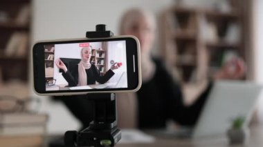 Blurred relaxed young muslim woman in casual wear sitting on workplace with modern laptop and meditating with closed eyes during recording video blog. Focus on modern smartphone fixed on tripod.