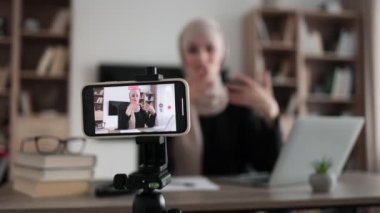 Blurred relaxed young muslim woman in casual wear sitting on workplace with modern laptop and meditating with closed eyes during recording video blog. Focus on modern smartphone fixed on tripod.