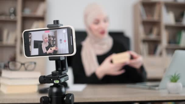 ギフトボックスをアンパックしながら カジュアルな服装でイスラム教徒の女性の携帯電話やスマートフォンでのヒジャーブ録画ビデオに焦点を当てます 女性ブロガーは ソーシャルネットワークで彼女の加入者と彼女の感情を共有します — ストック動画