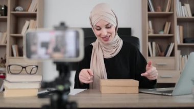 Pleasant muslim woman filming video on modern phone camera while opening parcel box with new smartphone. Concept of people, technology and blogging.