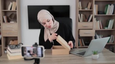 Pleasant muslim woman filming video on modern phone camera while opening parcel box with new smartphone. Concept of people, technology and blogging.