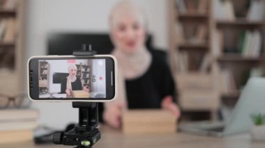 Focus on smart phone screen, pleasant arab woman filming video on modern phone camera while opening parcel box with new smartphone. Concept of people, technology and blogging.