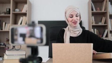 Pleasant arab woman filming video on modern phone camera while opening parcel box with new wireless laptop. Concept of people, technology and blogging.