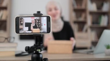 Pleasant arab woman filming video on modern phone camera while opening parcel box with new wireless headphones. Concept of people, technology and blogging.