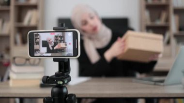 Focus on smart phone screen, pleasant arab woman filming video on modern phone camera while opening parcel box. Concept of people, technology and blogging.