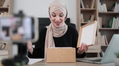 Arab woman recording video on phone camera while unpacking box with new wireless headphones. Female influencer sharing with subscribers her positive feedback about new order.