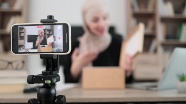 Pleasant arab woman filming video on modern phone camera while opening parcel box with new wireless headphones. Concept of people, technology and blogging.