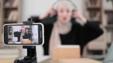Pleasant arab woman filming video on modern phone camera while opening parcel box with new wireless headphones. Concept of people, technology and blogging.