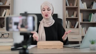 Muslim woman recording video on phone camera while unpacking box with new wireless tablet. Female influencer sharing with subscribers her positive feedback about new order.
