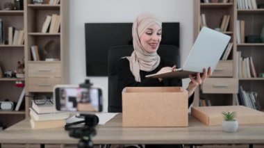 Pleasant arab woman filming video on modern phone camera while opening parcel box with new wireless laptop. Concept of people, technology and blogging.