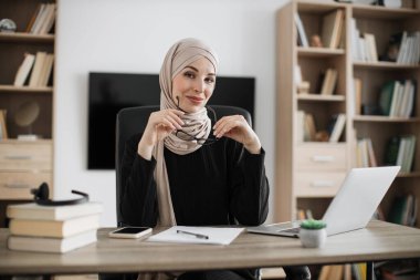 Confident smiling positive muslim woman in formal wear and hijab holding eyeglasses sitting at office desk. Company worker with headscarf looking at camera. Concept of people and business.