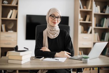 Smiling muslim woman in eyeglasses and hijab sitting at desk with books and notes. Female tutor working at home during distance learning.