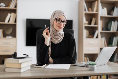 Smiling muslim woman in eyeglasses and hijab sitting at desk with books and notes. Female tutor working at home during distance learning.