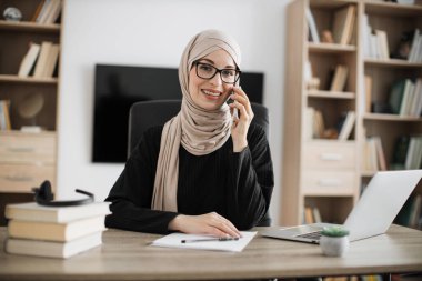 Muslim business woman having mobile conversation while sitting at table and working on wireless laptop. Working process at office of young arab girl in hijab.