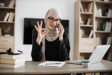 Muslim business woman having mobile conversation while sitting at table and working on wireless laptop showing sign ok. Working process at office of young arab girl in hijab.