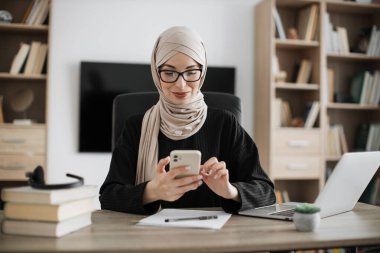 Beautiful young concentrated hardworking muslim businesswoman, it specialist or manager in hijab typing message on modern smartphone and working on laptop computer in office.