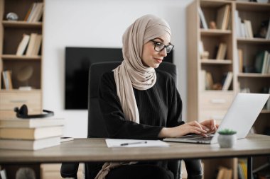 Side view, portrait of young smiling experienced smart stylish muslim manager in eyeglass, businesswoman in hijab sitting at table, using laptop at modern office.