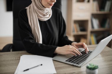 Cropped view, portrait of young smiling experienced smart stylish muslim manager in eyeglass, businesswoman in hijab sitting at table, using laptop at modern office.